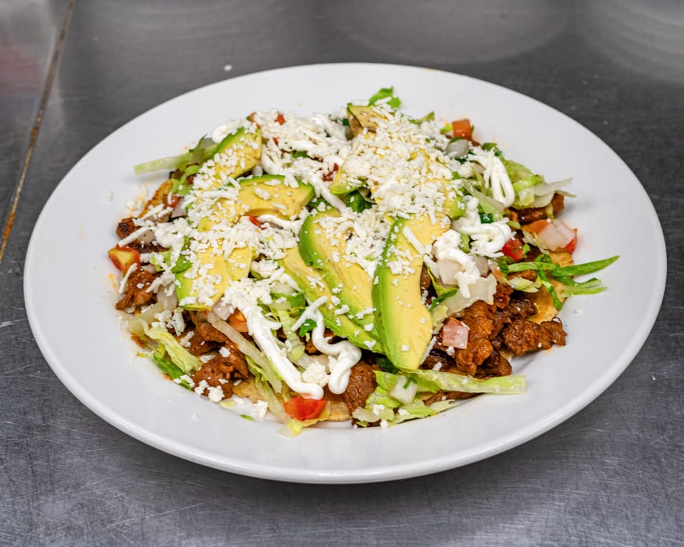 A white plate holds a Mexican dish with meat, lettuce, avocado slices, cheese, and crema.