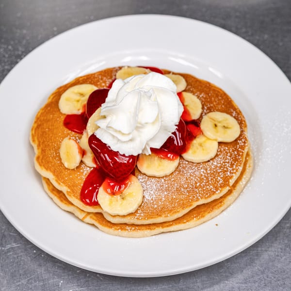 Fluffy pancakes topped with whipped cream, strawberry syrup, banana slices, and powdered sugar.