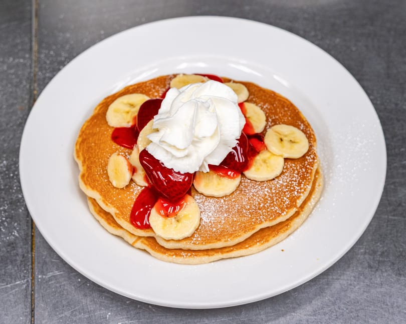 Fluffy pancakes topped with whipped cream, strawberry syrup, banana slices, and powdered sugar.