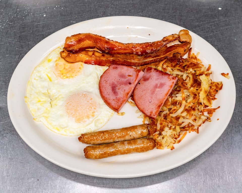 A full breakfast plate with two fried eggs, bacon, ham, hash browns, and two sausage links.