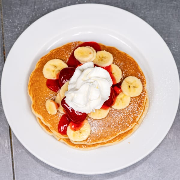 Fluffy pancakes topped with sliced bananas, strawberry syrup, and whipped cream on a white plate.