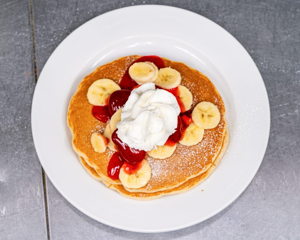Fluffy pancakes topped with sliced bananas, strawberry syrup, and whipped cream on a white plate.
