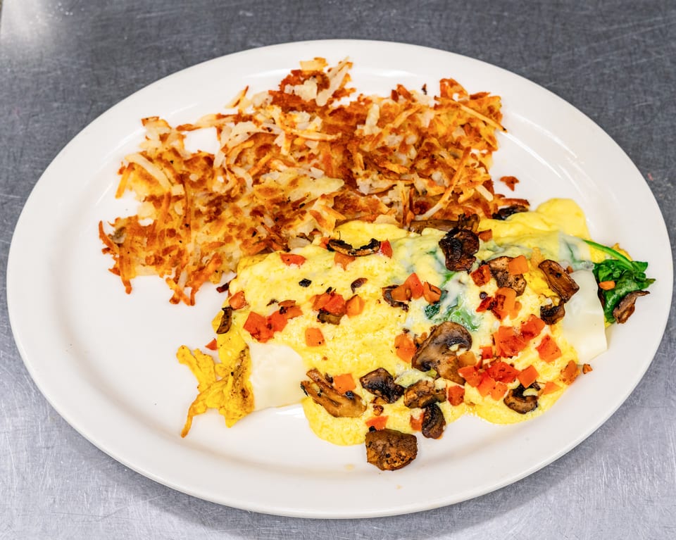 A white plate holds a golden-brown omelet with mushrooms, spinach, and diced tomatoes, alongside hash browns.
