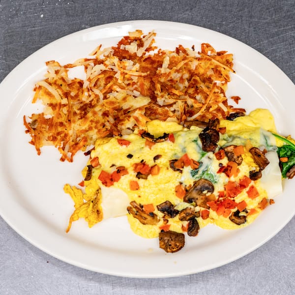 A white plate holds a fluffy omelet with mushrooms, tomatoes, and spinach, alongside crispy hash browns.