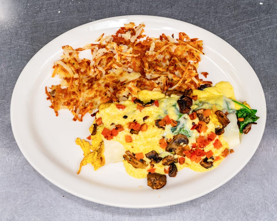 A white plate holds a fluffy omelet with mushrooms, tomatoes, and spinach, alongside crispy hash browns.