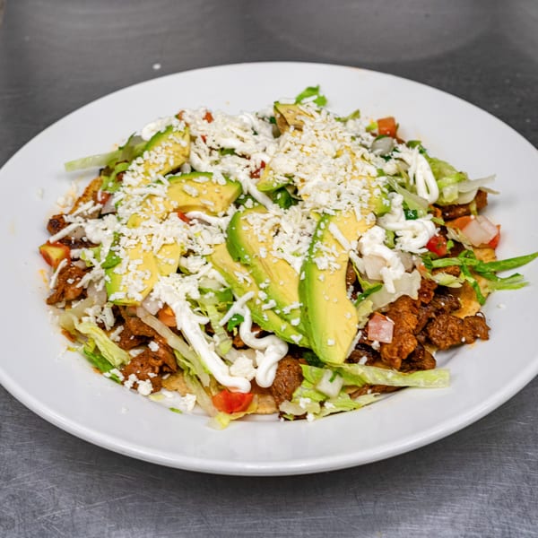 A white plate holds a Mexican dish with meat, lettuce, avocado slices, cheese, and sour cream.