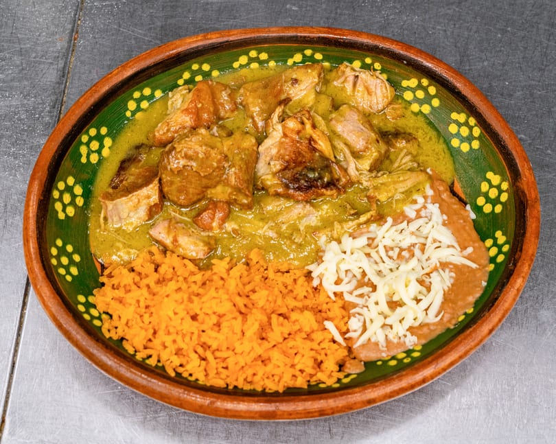 Pork in green chile sauce with orange rice and refried beans topped with cheese in a decorative bowl.
